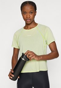 Αθλητική μπλούζα λαχανί με κοντά μανίκια και ανακλαστικές λεπτομέρειες, φορεμένη με μαύρα κολάν. Το άτομο κρατά ένα μαύρο μπουκάλι νερού.