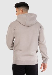 Man die een lichtgrijze hoodie draagt, gezien van achteren, met zwarte broek en een kort kapsel tegen een effen achtergrond.