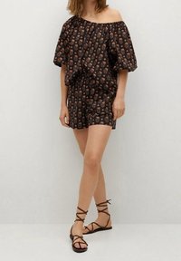 Sort off-the-shoulder romper med korte ærmer og blomsterprint i orange. Sammen med brune sandaletter med stropper, der har flade såler.