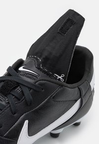 Scarpa da calcio in pelle nera con accenti bianchi, caratterizzata da una superficie testurizzata, dettagli cuciti e un design slip-on con una linguetta in velcro.