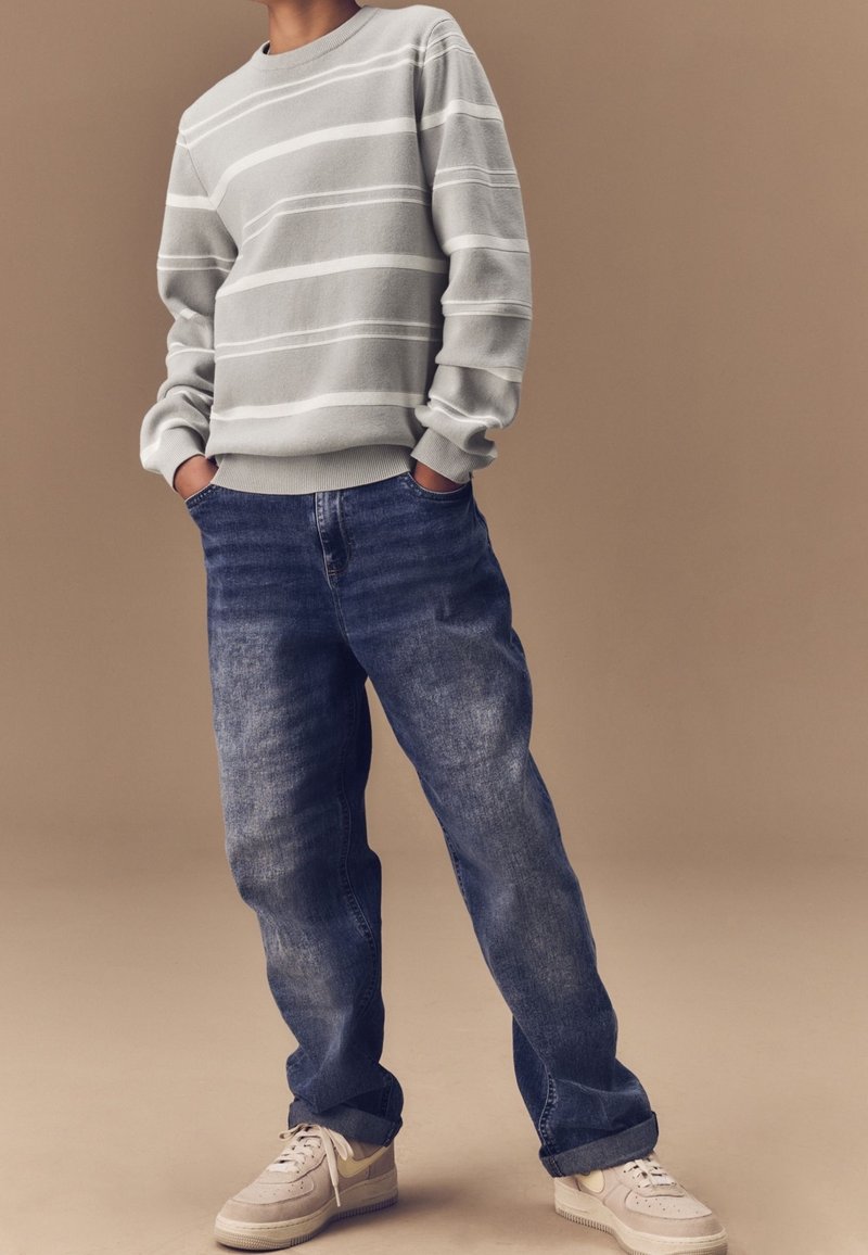 Sweatshirt à rayures grises avec un col rond, associé à un jean bleu ample. Les chaussures arborent une palette de couleurs grise clair et beige.