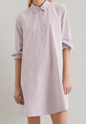 Femme portant une robe-chemise ample à manches longues avec des rayures verticales lavande, blanches et beige et des manches retroussées.