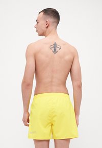 Ryškiai geltonos maudymosi kelnaitės su elastinga juosmena, kurių kairėje pusėje matomas logotipo detalė. Modelis matomas iš nugaros, kurioje atsiskleidžia tatuiruotė.