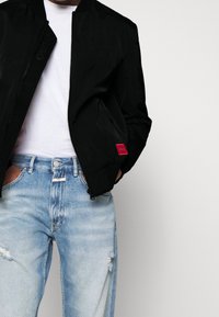 Veste bomber noire avec fermeture éclair à l'avant, col côtelé et un écusson de logo rouge. Portée sur un t-shirt blanc avec un jean bleu clair déchiré.