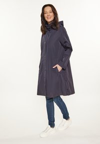 Impermeabile blu navy con cappuccio, bottoni a pressione e tasche laterali, caratterizzato da un taglio dritto e una texture liscia. Abbinato a jeans blu e sneakers bianche.