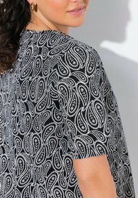 Černobílá látka s paisley vzorem, hladkou texturou, krátkými rukávy a kulatým výstřihem, vystavená na rameni modelky.