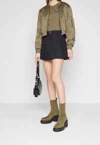 Chaqueta bomber de olivo, top verde ajustado, mini falda negra con pliegues, bolso negro y botines de olivo con suela gruesa.