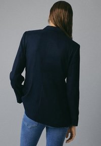 Persona con capelli lunghi che indossa un blazer blu scuro e jeans blu, in piedi con la schiena rivolta verso la telecamera su uno sfondo grigio uniforme.