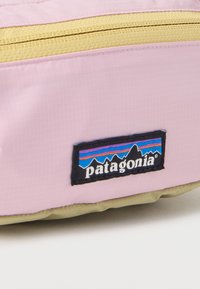 Κοντινό πλάνο μιας ροζ και μπεζ τσάντας Patagonia με κίτρινο φερμουάρ και μαύρο λογότυπο Patagonia που απεικονίζει ένα σχέδιο βουνού.