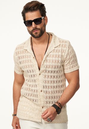 Beige, knitted short-sleeve shirt with a diamond pattern, button closure, and a collar; paired with dark sunglasses and layered bracelets.