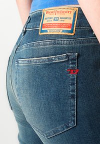 Blue denim jeans with a rear pocket and a Diesel Industry leather brand patch sewn above the pocket on the waistband.