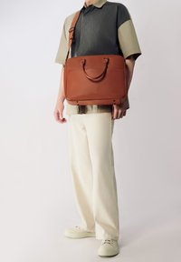 Bolsa de mensajero de cuero marrón con asas dobles y correa de hombro ajustable, textura suave, hardware minimalista, con un bolsillo frontal.