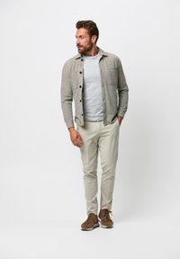 Camicia grigia con bottoni, dotata di due tasche sul petto, indossata sopra un maglione grigio chiaro a girocollo. I pantaloni beige e le sneakers marroni completano l'outfit.