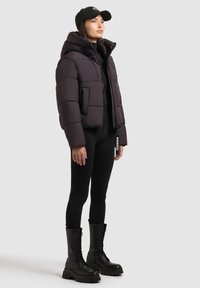 Puffermantel i mørk lilla med høj krave, der har en frontlomme og lynlås. Sorte leggings og kraftige sorte snørebåndsstøvler fuldender looket.