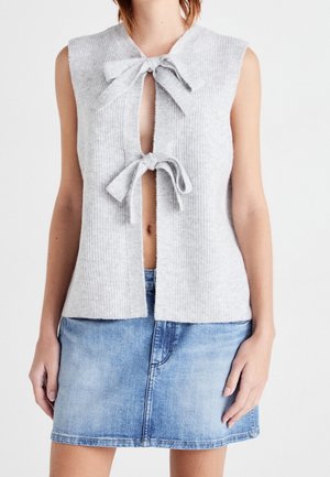 Haut tricoté gris clair sans manches avec deux nœuds à l'avant sur une mini-jupe en denim bleu porté par une personne debout sur fond blanc.