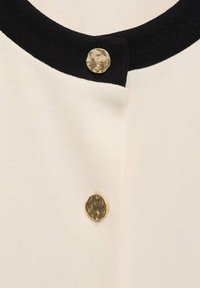 Stickad cardigan med en krämfärgad kropp och en svart ribbad halsringning. Två strukturerade guldknappar pryder knäppningen fram.