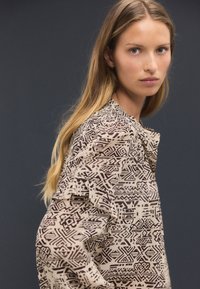 Beige patterned blouse featuring a tribal print in dark brown, with ruffled sleeves and a rounded neckline, made of lightweight fabric.