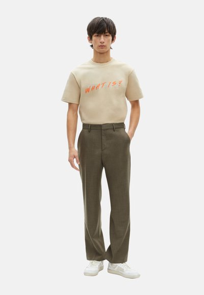 Camiseta de algodón beige con texto "¿QUÉ ES?" en naranja, combinada con unos pantalones ajustados en verde oliva claro y unas zapatillas blancas.