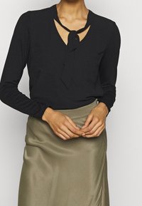 Svart långärmad topp med V-ringning och knytning vid kragen, kombinerad med en silkeslen olivgrön kjol som visar en mjuk textur.