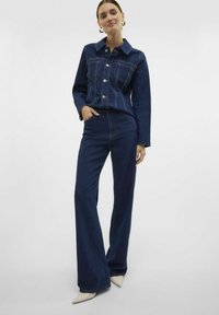 Denim jakna v temno modri barvi z dvema žepoma na prsih in srebrnimi gumbi, v kombinaciji z ujemajočimi se hlačami z visokim pasom in širokimi nogami, zaključeno s koničastimi bež čevlji.