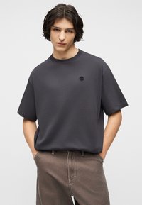 Νεαρό άτομο με μαύρα μεσαίου μήκους μαλλιά, που φοράει χαλαρό σκούρο γκρι Τ-shirt με μικρό λογότυπο και καφέ παντελόνι, με τα χέρια στις τσέπες.