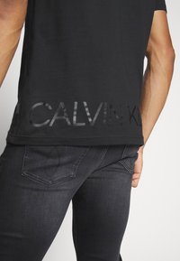 Černé bavlněné tričko s lesklým logem "CALVIN KLEIN" na spodním lemu. Upnutý střih s krátkými rukávy, zkombinované s tmavými denimovými džíny.