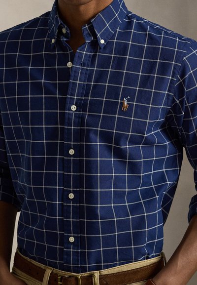 Navy blue button-down shirt with white grid pattern and a small embroidered logo. Cotton fabric with a smooth texture. Includes white buttons.