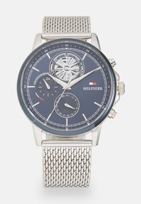 Orologio con cinturino a maglia argentata e quadrante blu scuro, dotato di sub-quadranti per data e ora, e un design del movimento visibile. Marca: Hilfiger.