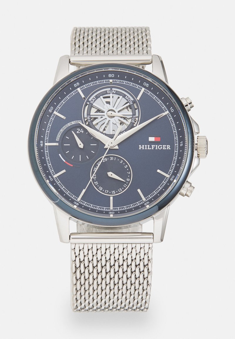 Orologio con cinturino a maglia argentata e quadrante blu scuro, dotato di sub-quadranti per data e ora, e un design del movimento visibile. Marca: Hilfiger.