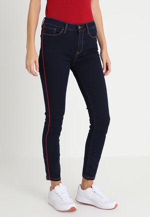 Vrouw draagt donkere skinny jeans met rode zijstreep, rood topje en witte sneakers, staand tegen een effen achtergrond.