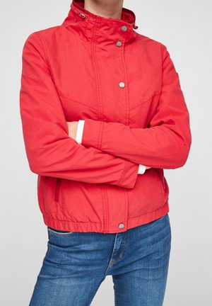 Personne portant une veste rouge vif avec des boutons-pression et un jean bleu, les bras croisés, sur un fond uni gris clair.
