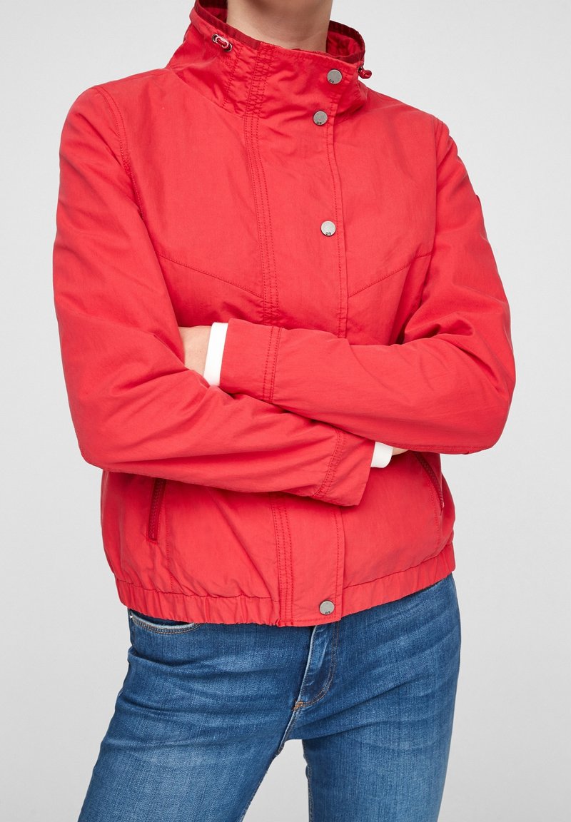 Personne portant une veste rouge vif avec des boutons-pression et un jean bleu, les bras croisés, sur un fond uni gris clair.
