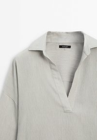 Chemise gris clair à fines rayures verticales, col ouvert et patte de boutonnage en V, avec l'étiquette de la marque visible à l'intérieur du col.