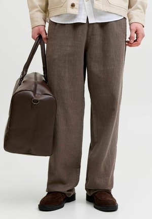 Homme portant un pantalon marron texturé et des chaussures marron foncé tenant un grand sac de voyage en cuir marron foncé par les poignées.