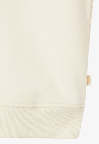 Materiał w kolorze kremowym z prążkowanym wykończeniem i małą beżową metką z pomarańczowym tekstem po prawej stronie.