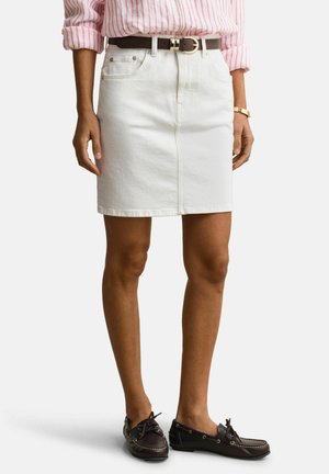 Frau trägt weißen Jeansrock mit Fronttaschen, braunem Gürtel, pink gestreiftem Hemd, goldener Uhr und dunkelbraunen Mokassins, steht vor weißem Hintergrund.