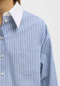 Chemise à rayures bleu clair avec col blanc, boutonnière frontale, et poche sur le côté gauche de la poitrine. Tissu doux avec une coupe décontractée et manches longues.