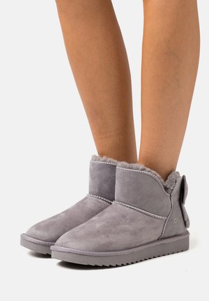 Gray suede ankle boots with a soft fur lining and a ribbon accent at the back, featuring a flat rubber sole and minimal stitching detail.