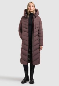 Lang, bordeauxpufferjakke med hætte, med chevron-quiltet design, frontlynlås og to sidelommer. Båret over en sort turtleneck.