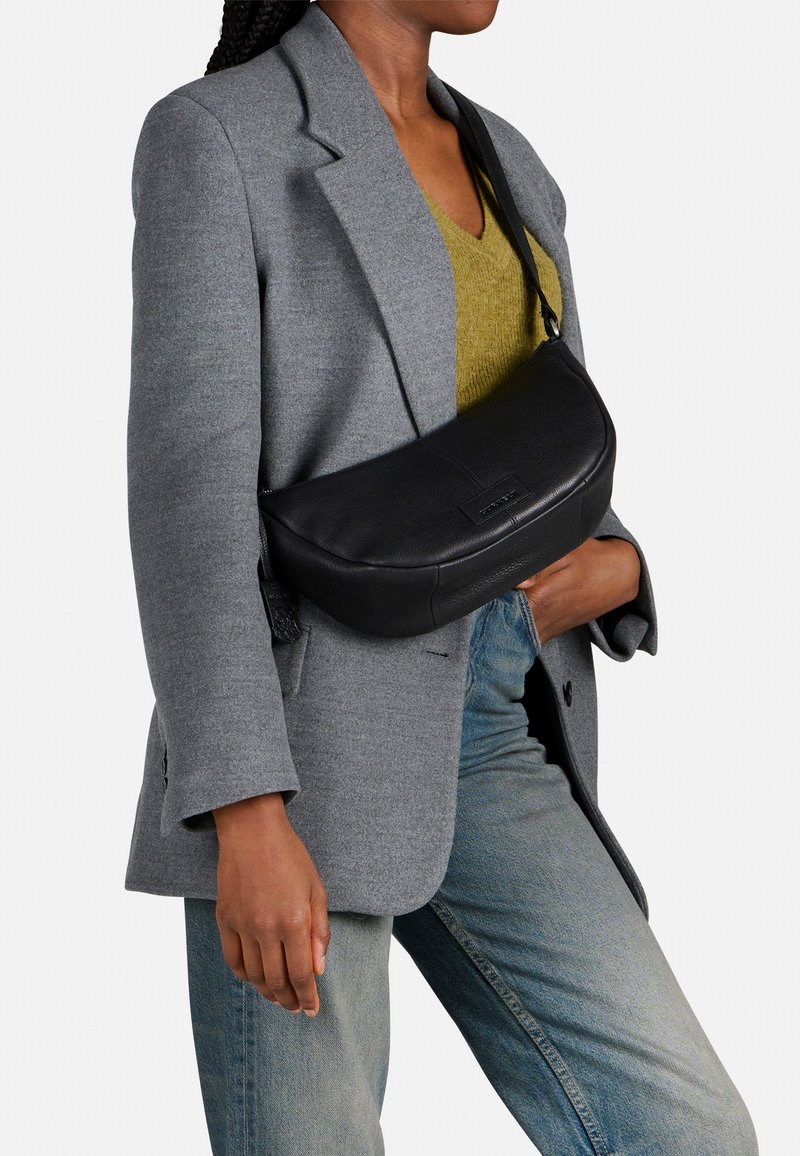 Borsa a spalla nera in pelle, forma curvata, superficie testurizzata, con una sottile tracolla. Abbinata a un blazer grigio e a un top verde.