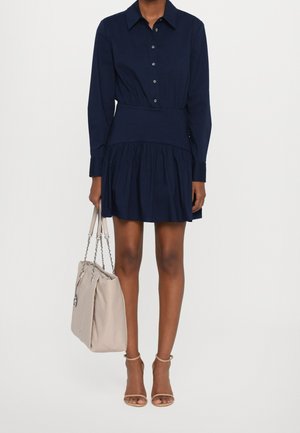 Femme portant une robe boutonnée à manches longues bleu marine tenant un sac à main beige, debout avec des pieds en escarpins à bride cheville couleur nude sur fond blanc.