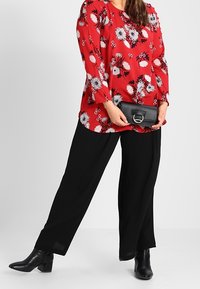 Rød blomsterbluse med sorte accenter, løs pasform, parret med sorte brede bukser og sorte stilethæle. Clutch med cirkulært hardware.