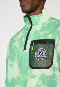 Πράσινο πουλόβερ με τεταρτημόριο φερμουάρ κα ι tie-dye σχέδιο, που διαθέτει διχτυωτή τσέπη με κυκλικό λογότυπο και μαύρες λεπτομέρειες, καθώς και ψηλό γιακά και κλείσιμο με φερμουάρ.