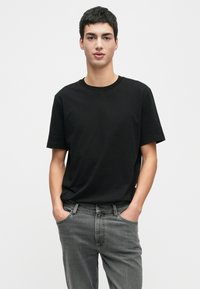 Jovem vestido com uma t-shirt preta simples e jeans cinzentos, de pé com as mãos nos bolsos contra um fundo branco liso.