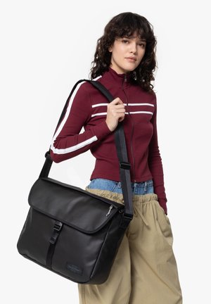 Black messenger bag with a flap closure and adjustable strap. Made of smooth material, featuring a front buckle and zippered pocket.