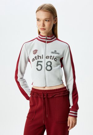 Cropet lynlåsjakke i lys grå med maroon ærmer og striber. Har atletiske grafikker og en tætsiddende design. Parret med matchende bukser.