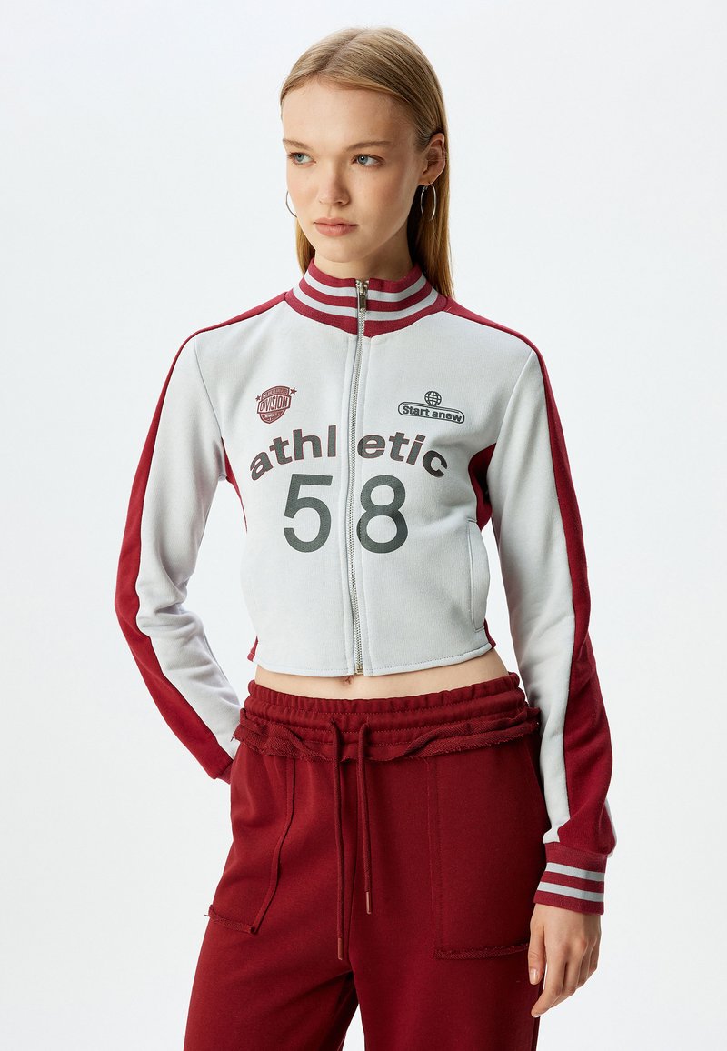 Cropet lynlåsjakke i lys grå med maroon ærmer og striber. Har atletiske grafikker og en tætsiddende design. Parret med matchende bukser.