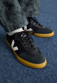 Melni un zaļi sporta apavi ar zamšādas un auduma materiāliem, ar dzeltenu gumijas zoli un lielu baltu logos sānos.
