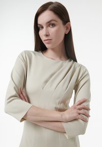 Vestido beige con escote redondo, mangas largas y pliegues verticales. Está hecho de tela suave y presenta una silueta ajustada sin adornos.