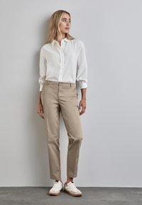Chinos beige con gamba dritta, chiusura a bottone e tasche laterali, abbinati a una camicia bianca con bottoni e sneakers bianche.